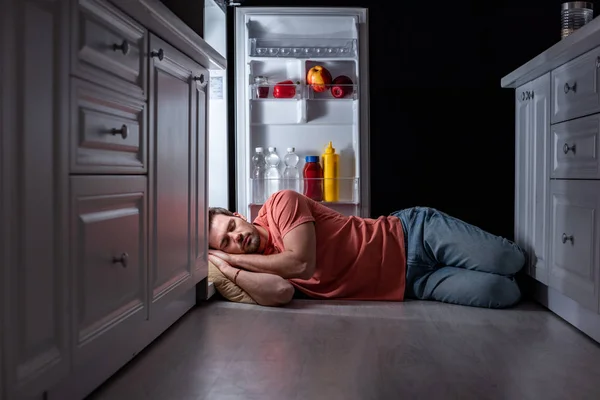 Exhausted man sleeping near open refrigerator on kitchen floor — Stock Photo