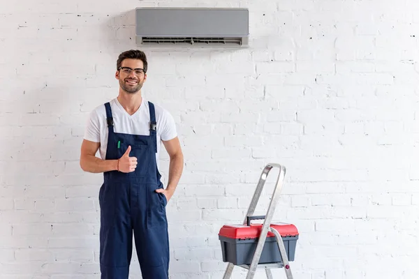 Reparador alegre mostrando el pulgar hacia arriba mientras está de pie bajo el aire acondicionado cerca de escalera y caja de herramientas - foto de stock