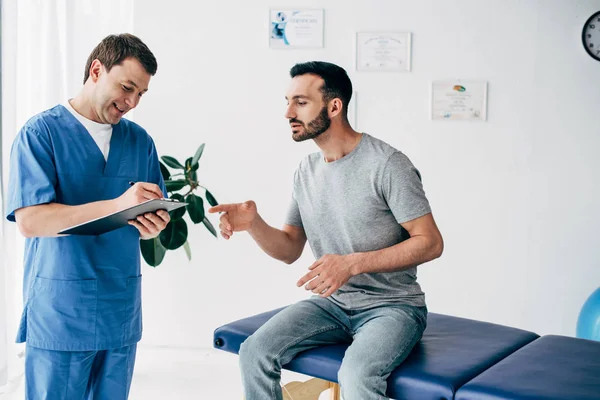 Patient zeigt mit dem Finger auf Klemmbrett, während lächelnder Arzt Rezept in Massageschrank in Klinik schreibt — Stockfoto