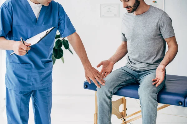 Vista ritagliata del medico che esamina il ginocchio del paziente e tiene appunti nell'armadietto dei massaggi in clinica — Foto stock