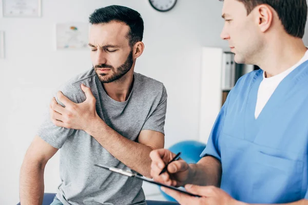 Selective focus of patient touching his shoulder and suffering from pain and doctor writing prescription in massage cabinet at clinic — Stock Photo