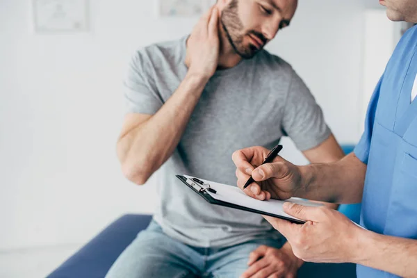 Selective focus of doctor writing on clipboard and suffering patient sitting on couch and touching neck in massage cabinet at clinic — Stock Photo