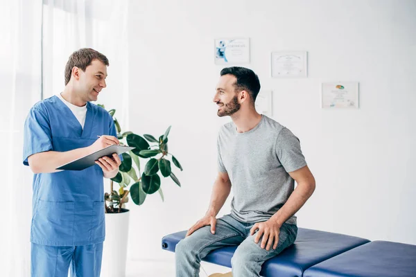 Усміхнений пацієнт сидить на дивані і розмовляє з лікарем в масажному кабінеті в клініці — стокове фото