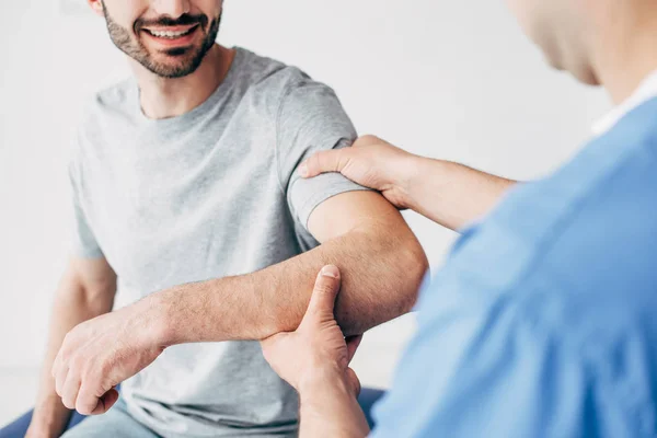 Selective focus of smiling patient sitting on couch and chiropractor examining patient shoulder in massage cabinet at clinic — Stock Photo