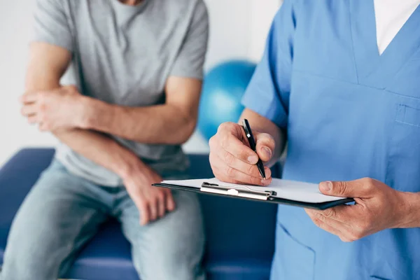 Cropped view of doctor writing on clipboard and patient touching arm in massage cabinet at clinic — Stock Photo