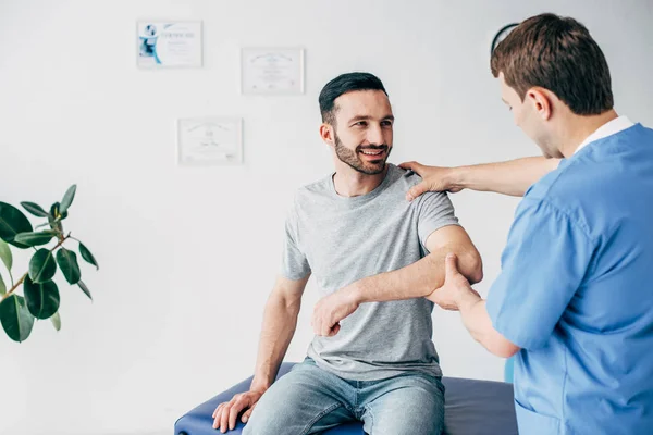 Paziente sorridente seduto sul divano e medico che esamina la spalla del paziente nell'armadietto dei massaggi in clinica — Foto stock