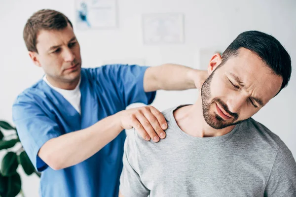 Selective focus of suffering man and doctor massaging man neck in massage cabinet at clinic — Stock Photo