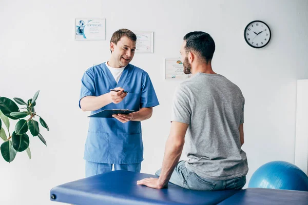 Fisioterapeuta sonriente con diagnóstico y gesto de pluma cerca del paciente en el hospital - foto de stock