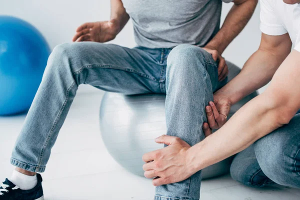 Cropped view of Physiotherapist massaging leg of patient sitting on fitness ball in hospital — Stock Photo