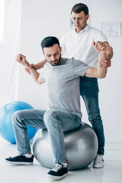Physiotherapeut streckt Arm einer hübschen Patientin auf Fitnessball im Krankenhaus — Stockfoto