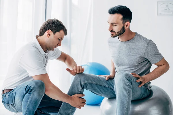 Physiotherapist massaging leg of handsome patient sitting on fitness ball in hospital — Stock Photo