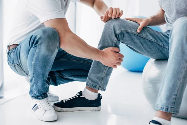 Vista ritagliata del fisioterapista massaggiare gamba del paziente seduto sulla palla fitness in ospedale — Foto stock