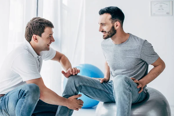 Physiothérapeute massant la jambe d'un patient souriant assis sur une balle de fitness à l'hôpital — Photo de stock