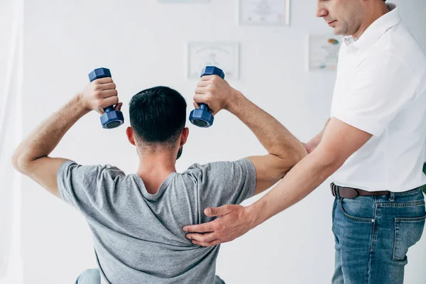 Chiropraktiker massiert Schulter von Mann mit Kurzhanteln im Krankenhaus — Stockfoto