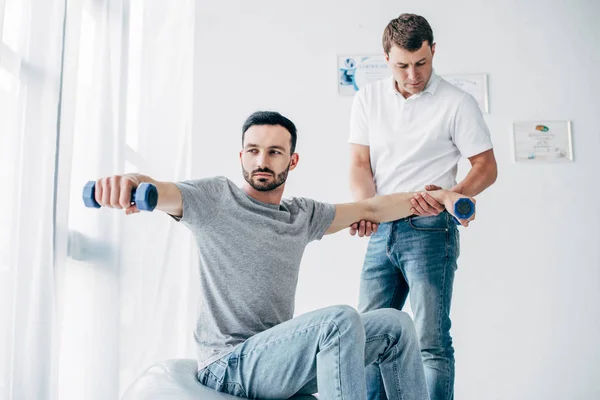 Chiropractor stretching arm of good-looking patient with dumbbells in hospital — Stock Photo