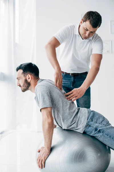 Chiropractor massaging back of handsome man lying on fitness ball — Stock Photo