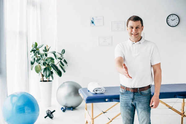 Lächelnder Chiropraktiker mit ausgestreckter Hand und Blick in die Kamera im Massageschrank — Stockfoto