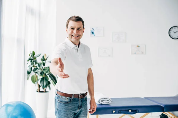 Lächelnder Chiropraktiker mit ausgestreckter Hand und Blick in die Kamera im Massageschrank — Stockfoto