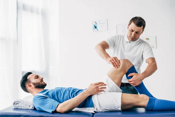 Fisioterapista massaggiare gamba di giocatore di calcio sdraiato sul lettino da massaggio in ospedale — Foto stock
