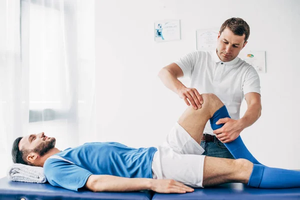 Bello fisioterapista massaggiare gamba di giocatore di calcio in ospedale — Foto stock