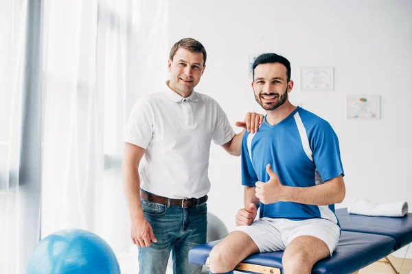 Smiling Physiotherapist near happy football player showing thumb up in hospital — Stock Photo