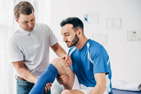 Fisioterapeuta masajeando pierna de guapo jugador de fútbol en el hospital - foto de stock