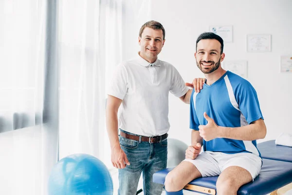 Smiling Physiotherapist near football player showing thumb up in hospital — Stock Photo