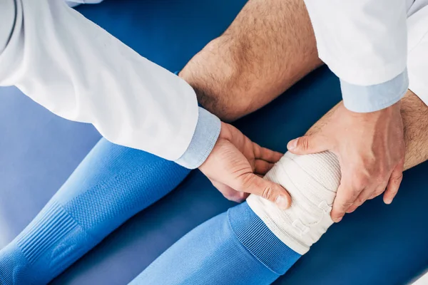 Vista recortada de fisioterapeuta poniendo vendaje elástico en la pierna del jugador de fútbol en la mesa de masaje - foto de stock