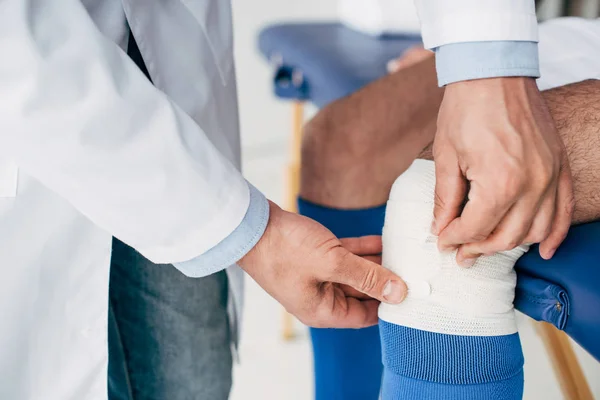 Частичный взгляд физиотерапевта в белом халате, накладывающего эластичный бинт на ногу футболиста — стоковое фото