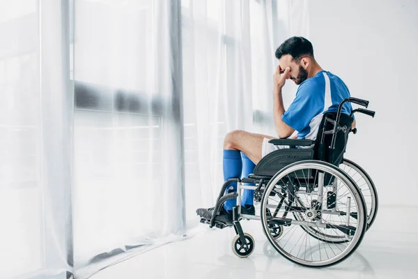 Расстроенный мужчина в футбольной форме сидит в инвалидной коляске в больнице возле окна — стоковое фото