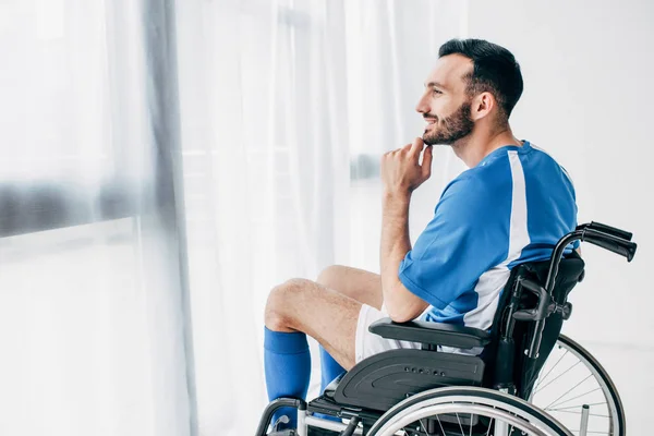 Hombre sonriente en uniforme de fútbol sentado en silla de ruedas y mirando a través de la ventana - foto de stock