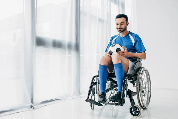 Hombre guapo en uniforme de fútbol sentado en silla de ruedas y sosteniendo pelota de fútbol - foto de stock