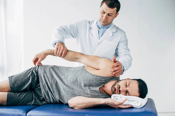 Chiropractor stretching arm of handsome patient lying on massage table in hospital — Stock Photo