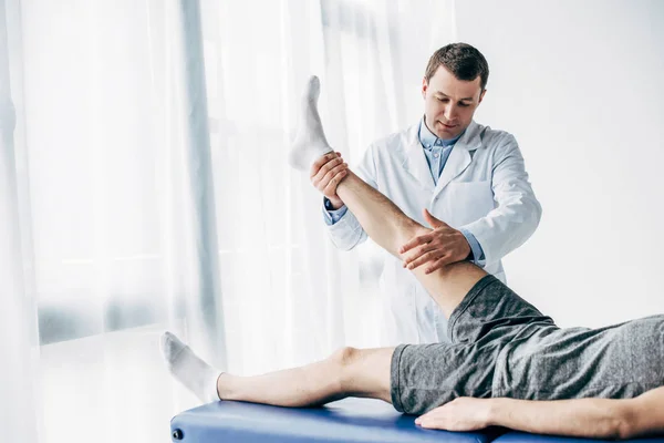 Handsome Physiotherapist stretching leg of patient on massage table in hospital — Stock Photo