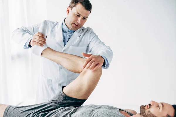 Handsome chiropractor stretching leg of patient in hospital — Stock Photo