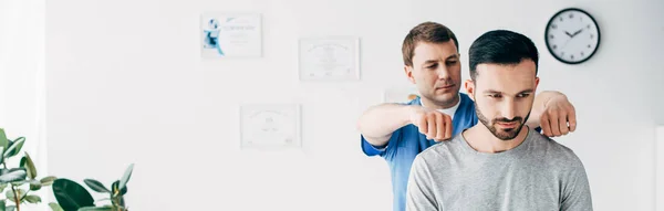 Panoramic shot of chiropractor massaging neck of handsome man in hospital — Stock Photo