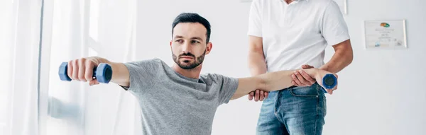 Panoramic shot of chiropractor stretching arm of handsome patient with dumbbells in hospital — Stock Photo