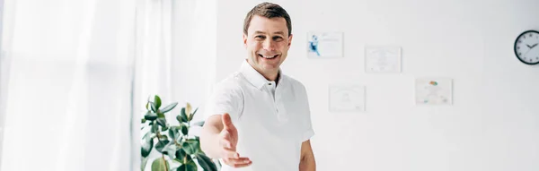 Panoramic shot of smiling chiropractor with outstretched hand looking at camera in hospital — Stock Photo