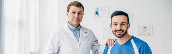 Panoramaaufnahme eines lächelnden Physiotherapeuten im weißen Kittel und einer Patientin, die im Krankenhaus in die Kamera schaut — Stockfoto