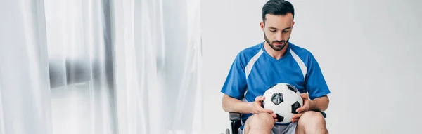 Tiro panorámico del hombre en uniforme de fútbol sentado en silla de ruedas y sosteniendo pelota de fútbol - foto de stock