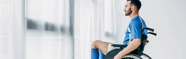 Tiro panorámico del hombre en uniforme de fútbol sentado en silla de ruedas cerca de la ventana y las cortinas - foto de stock