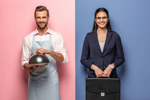 Happy man in apron with Serving Tray and businesswoman with briefcase on blue and pink — Stock Photo