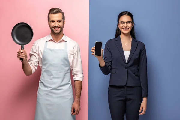 Чоловік в фартусі тримає сковороду, в той час як бізнес-леді тримає смартфон з порожнім екраном на синьому і рожевому — стокове фото