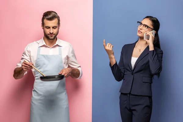Чоловік в фартусі тримає сковороду, поки бізнес-леді розмовляє на смартфоні на синьому і рожевому — стокове фото