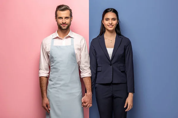 Hombre sonriente en delantal y mujer de negocios tomados de la mano en azul y rosa - foto de stock