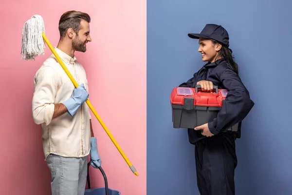 Hombre en guantes de goma con fregona y mujer en uniforme de trabajador de la construcción con caja de herramientas en azul y rosa - foto de stock