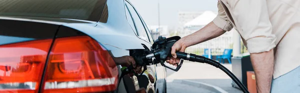 Panoramic shot of man holding fuel pump and refueling black car — Stock Photo