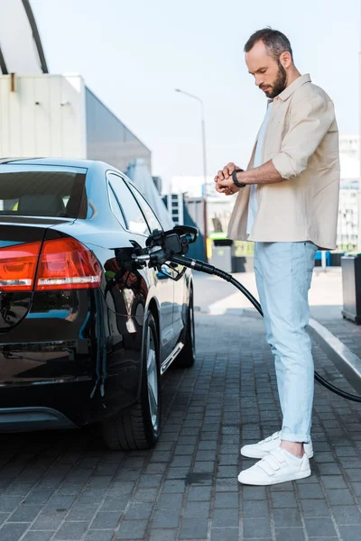 Handsome man standing and looking at watch while refueling black car at gas station — Stock Photo