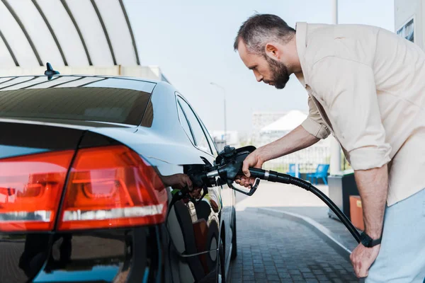 Bearded man holding fuel pump and refueling black car at gas station — Stock Photo