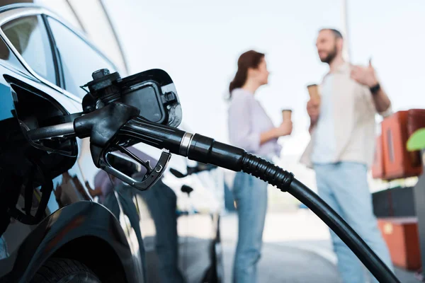 Selective focus of black automobile refueling with benzine near man and woman on gas station — Stock Photo